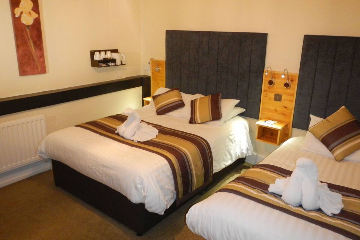 A hotel room with two beds, each with striped bedding and towels. There is a nightstand with a lamp and a small table with a cup and a book.