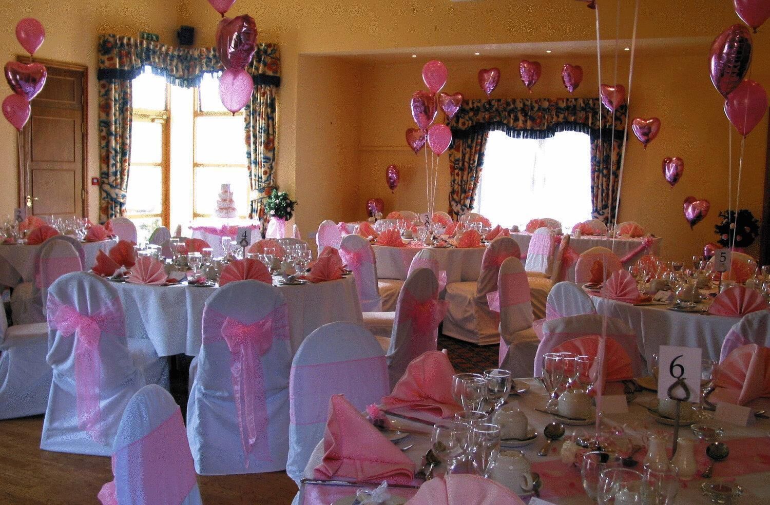 Un salón de banquetes con mesas redondas, manteles rosa y blancos, y sillas con cintas rosas.