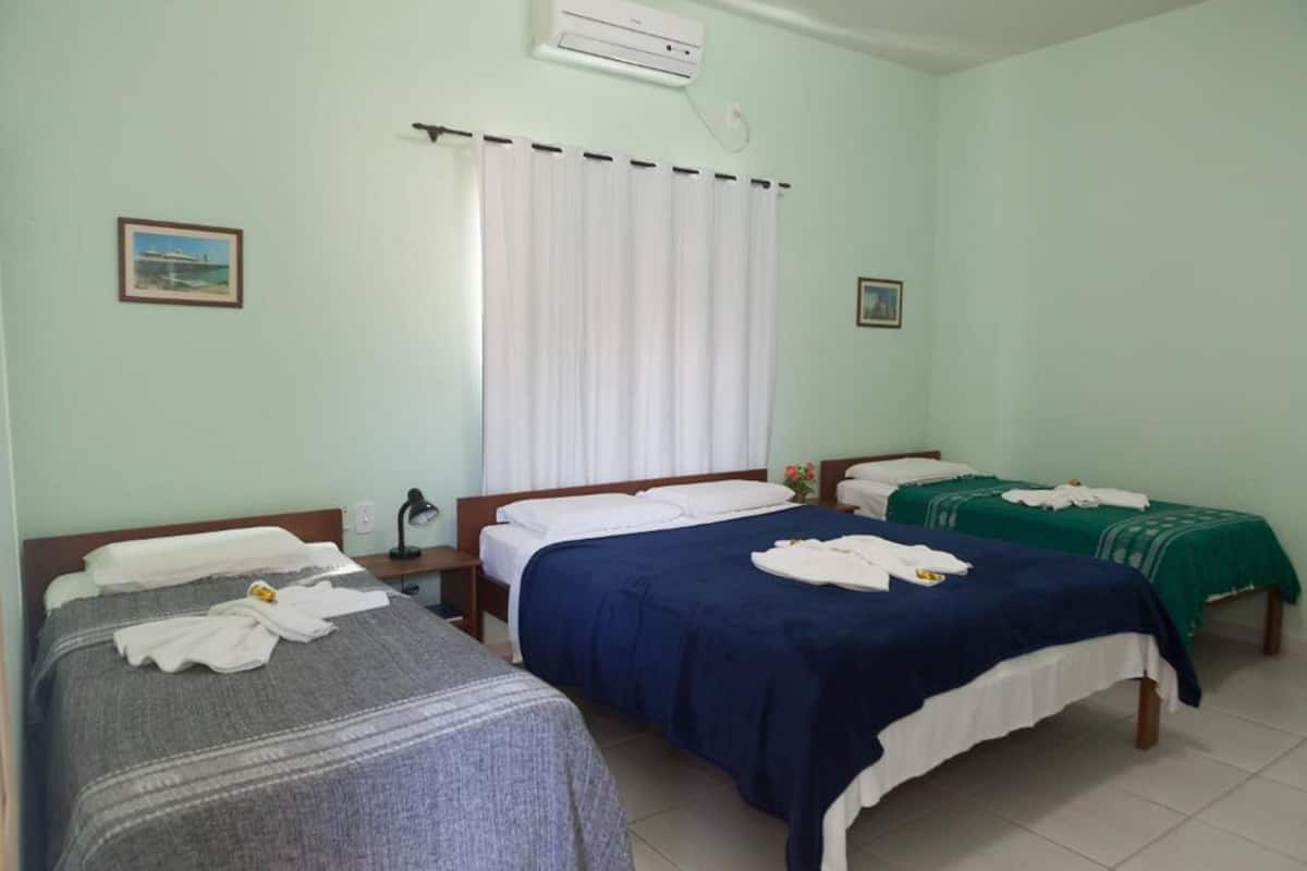 A hotel room with two single beds, a large window with white curtains, and a wall-mounted air conditioner.