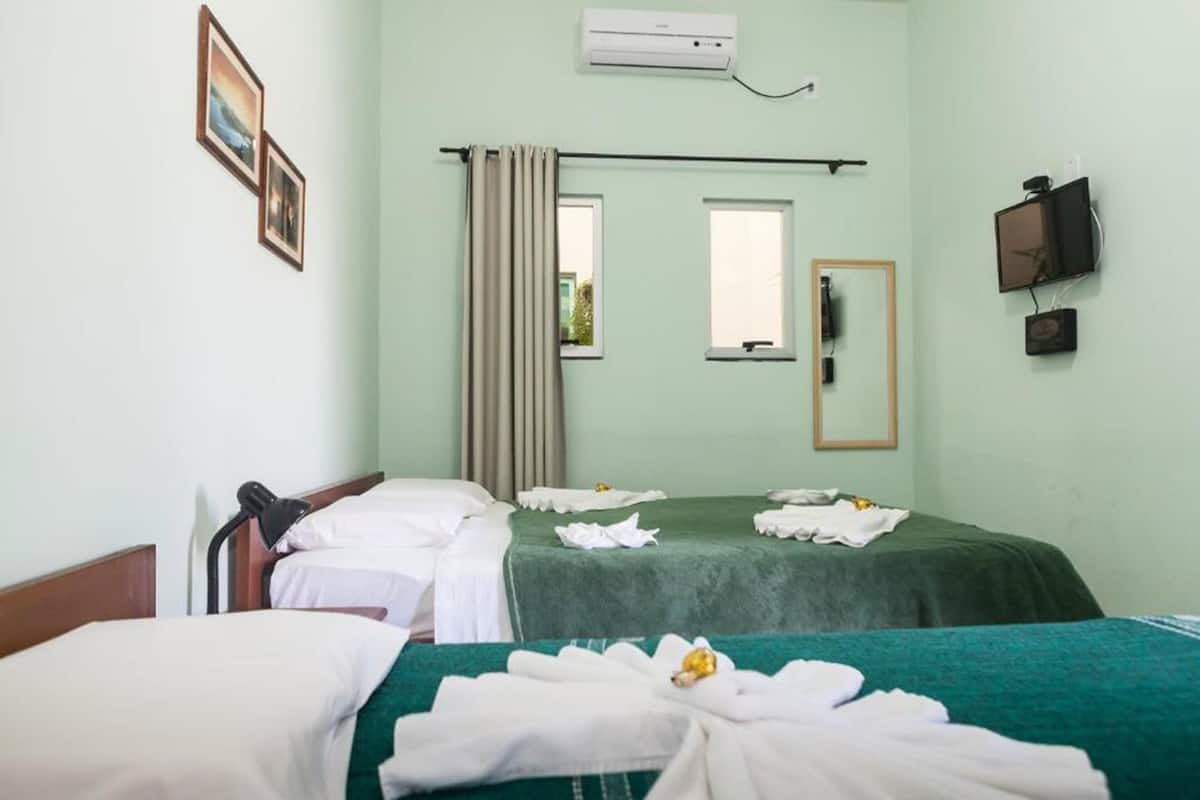 A hotel room with two beds, a window with curtains, a mounted TV, and an air conditioning unit.