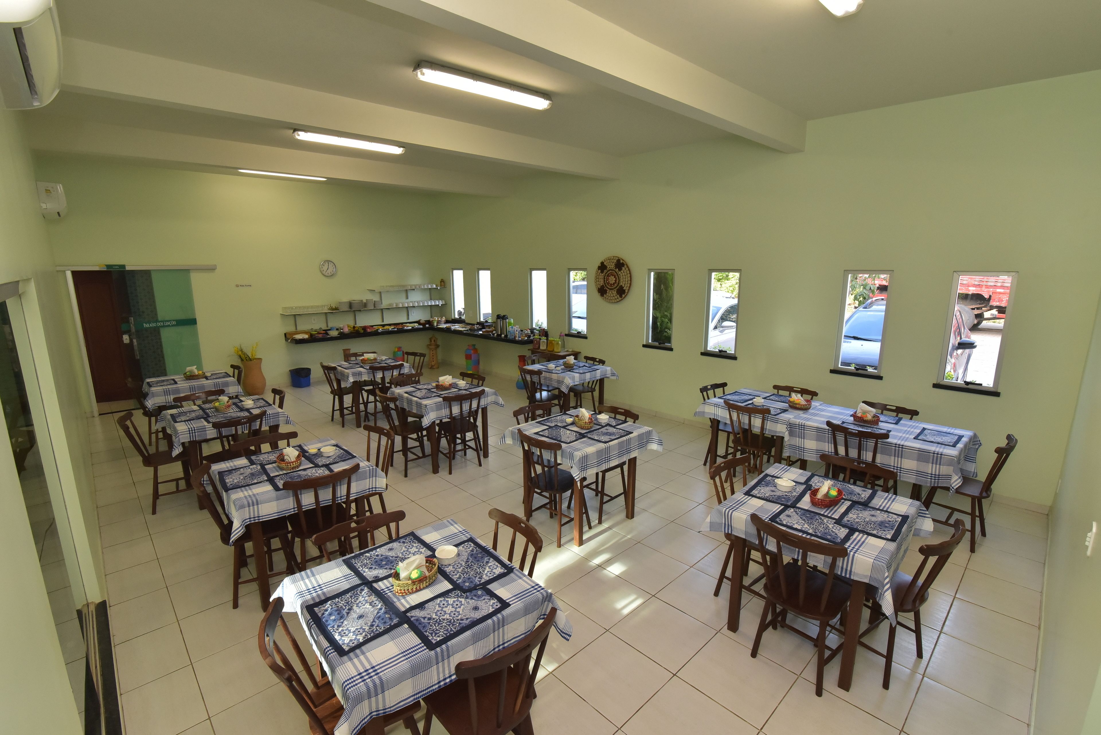 A dining area with multiple tables and chairs, a kitchen area with shelves and appliances, and a view of the outside through windows.