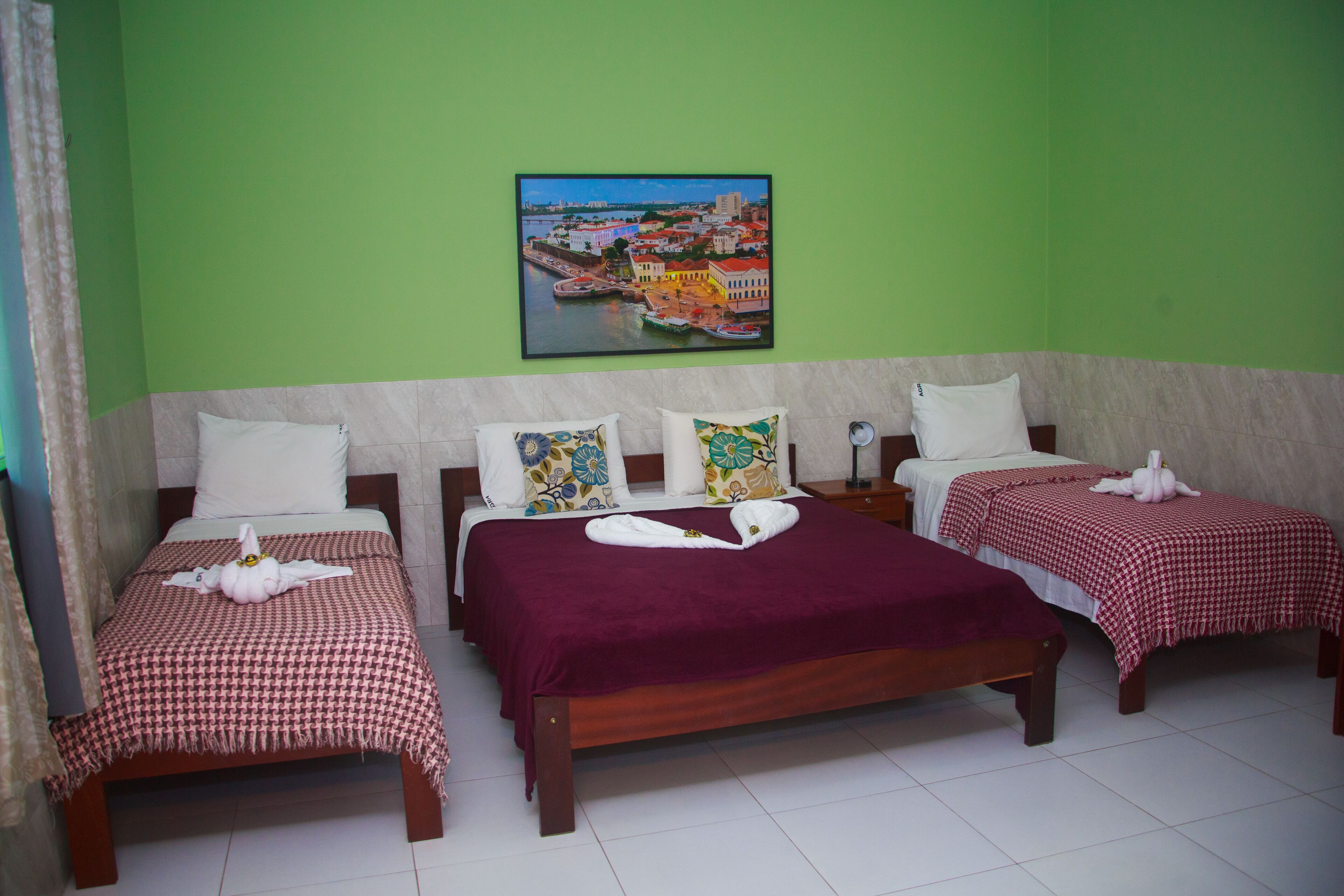 Habitación de hotel con tres camas, cada una con colcha y almohadas a cuadros, un cuadro enmarcados con una vista urbana y una vista de la ciudad por la ventana.
