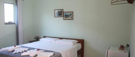 A hotel room with a bed, a small table with a chair, a window with curtains, and a wall-mounted shelf with a few items.
