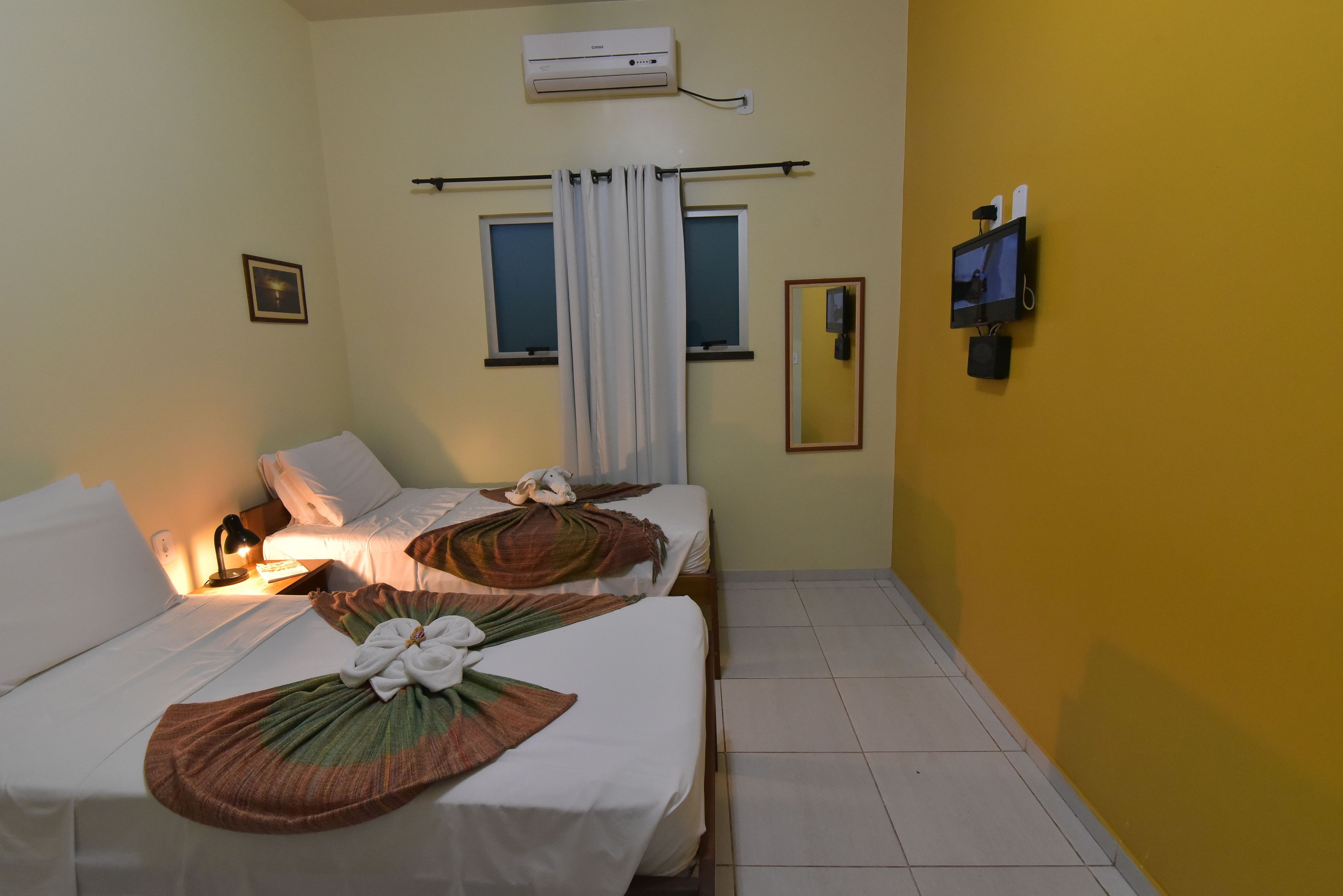 A hotel room with two beds, a flat-screen TV mounted on the wall, an air conditioning unit above the beds, and a window with curtains.