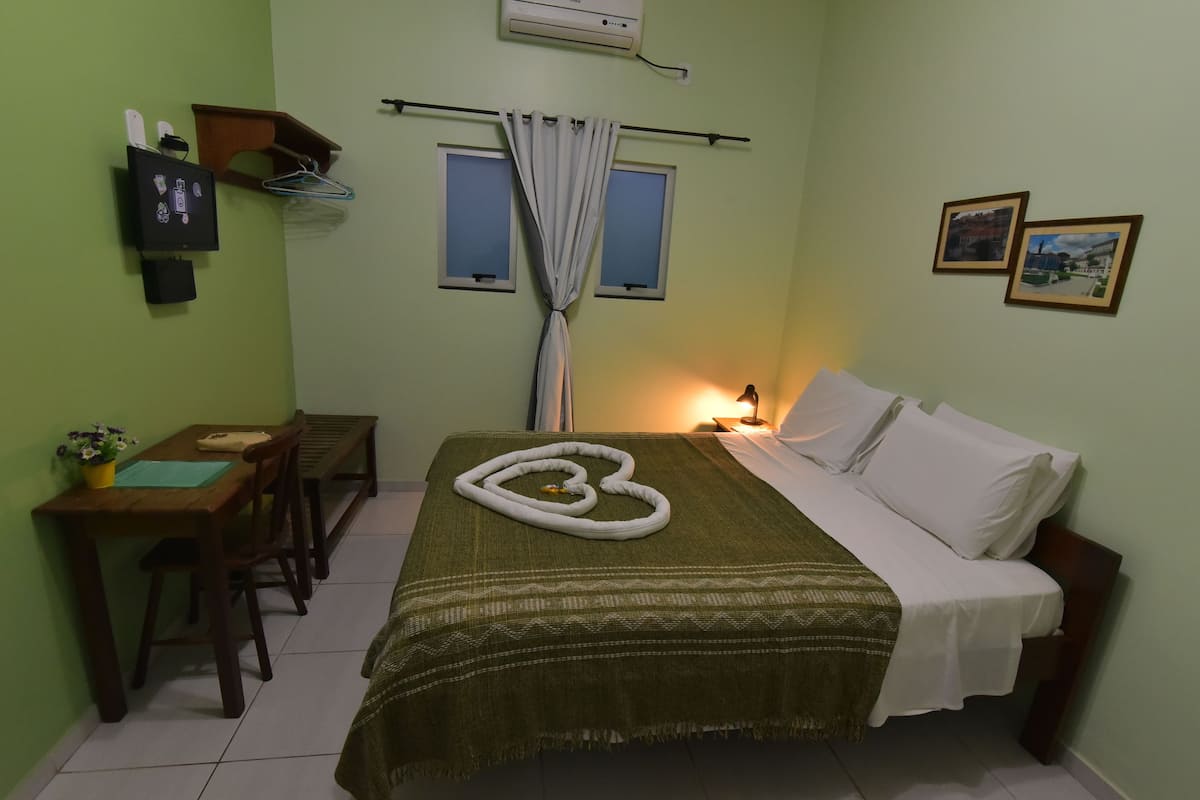 A small, well-lit room with a bed, a desk, and a chair. There is a window with curtains, a wall-mounted air conditioner, and a wall-mounted TV.