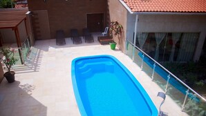 Una piscina rectangular con barandilla de cristal en un lado, rodeada por un patio con baldosas, sillas de descanso y plantas en macetas.