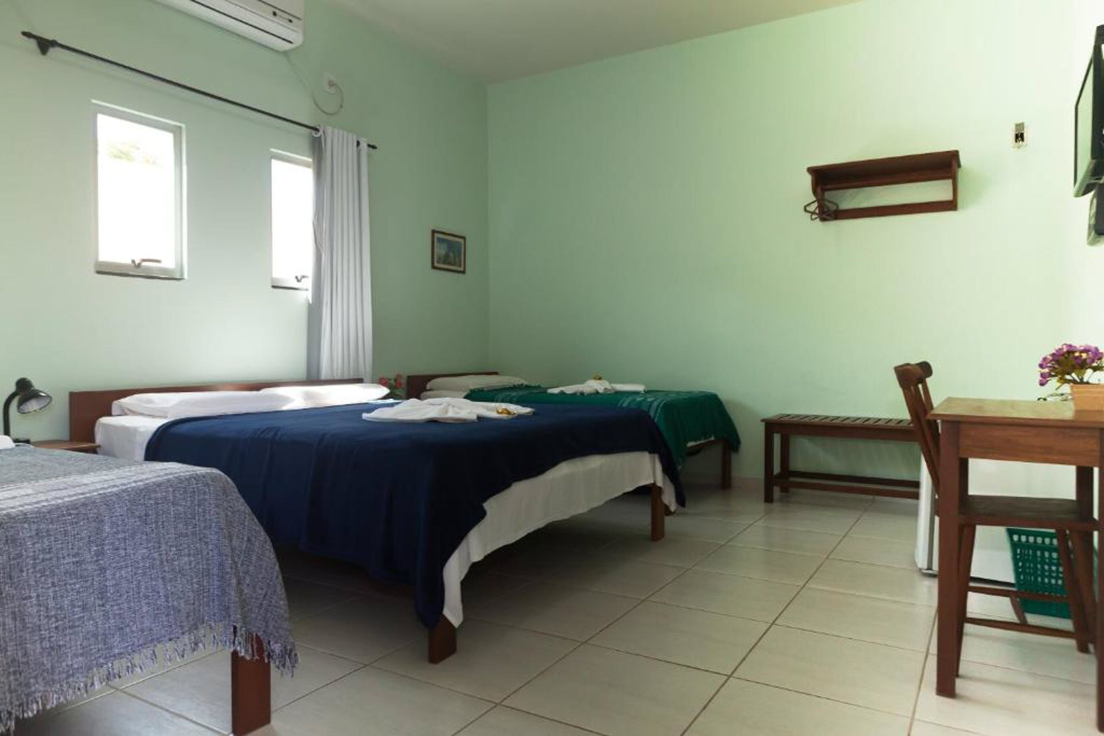 Una habitación con dos camas, una mesita, una silla, un ventanal y una estantería en la pared.