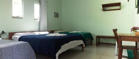 Una habitación con dos camas, una mesita, una silla, un ventanal y una estantería en la pared.