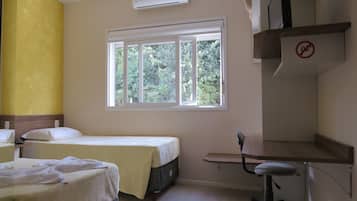 Quarto de hotel com duas camas, uma escrivaninha, uma cadeira, ar condicionado e janela com vista para árvores.