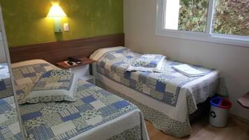 A room with two beds, a nightstand, a lamp, a window with a view of greenery, and a trash bin.