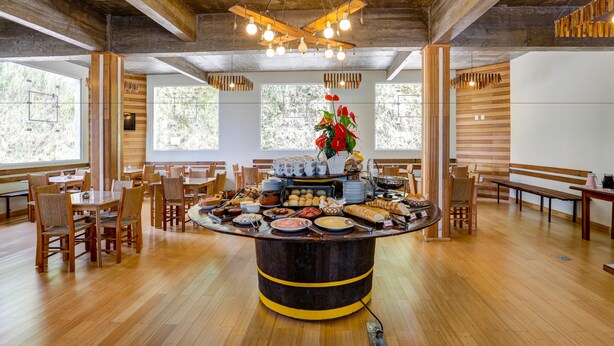 A dining area with a central buffet table surrounded by wooden chairs and tables, large windows, and a wooden ceiling. - Adoro Hotel (Farroupilha)