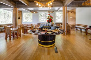Un espace repas avec une table de buffet centrale, entourée de chaises et de tables en bois, de grandes fenêtres et un plafond en bois.