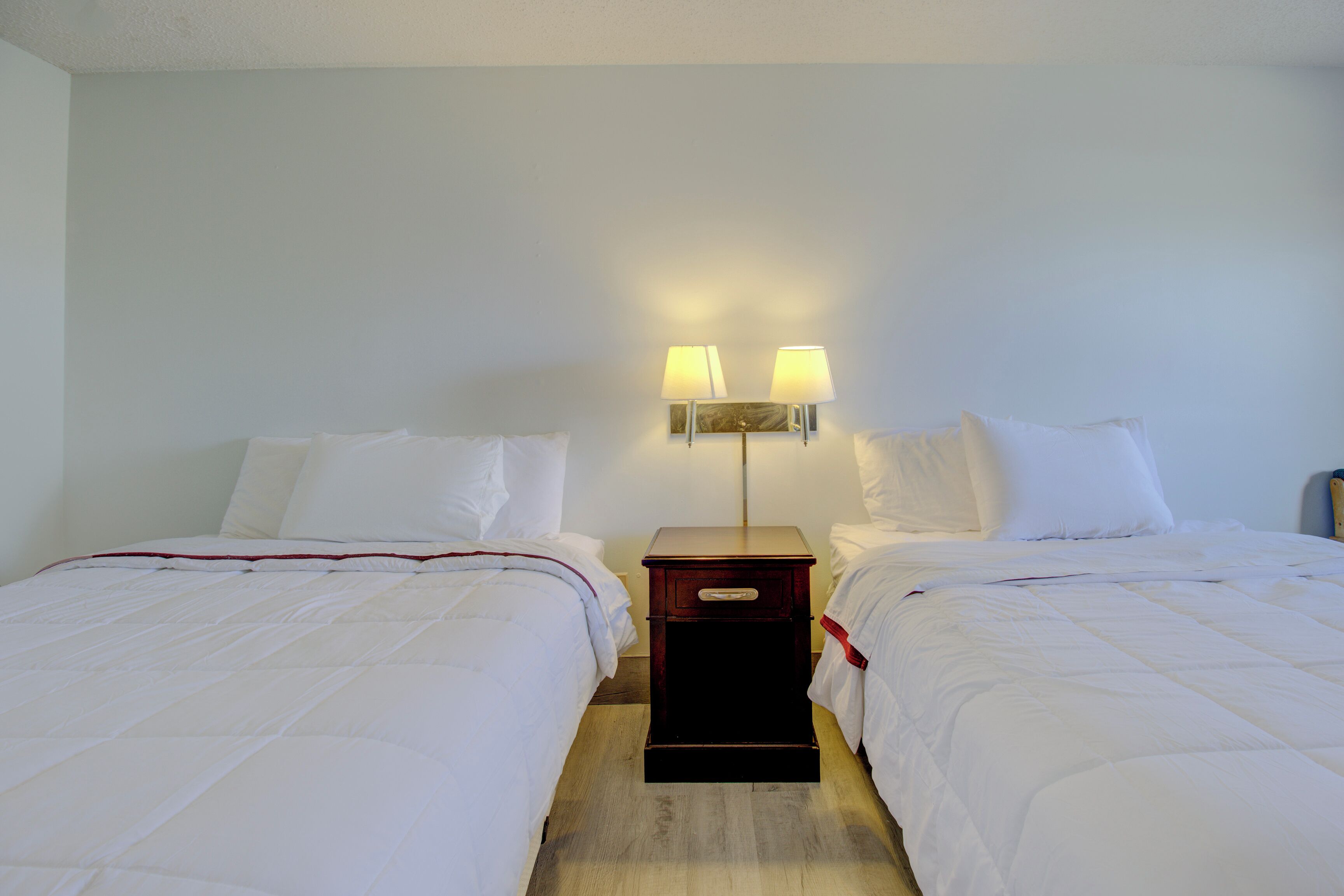 Una habitación de hotel con dos camas individuales, una mesita de noche con lámpara y suelo de madera.