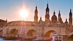 A stone bridge with multiple arches spanning a body of water, leading to a historic building with several towers and domes. - Hostal Ávila (Zaragoza)