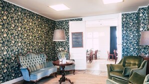 Um quarto com papel de parede floral, um sofá azul, uma poltrona verde, uma mesinha de cabeceira com uma luminária e uma porta que dá para outro cômodo.