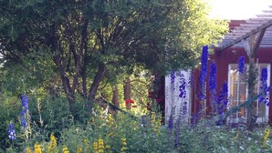 Un jardín con diversas plantas y flores, una pérgola de madera y un edificio rojo con ventanas.