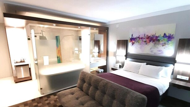 A hotel room with a large bed, a sofa, a bathroom with a glass shower, and a painting on the wall. - Palm Swift Luxury Accommodation (Brits)