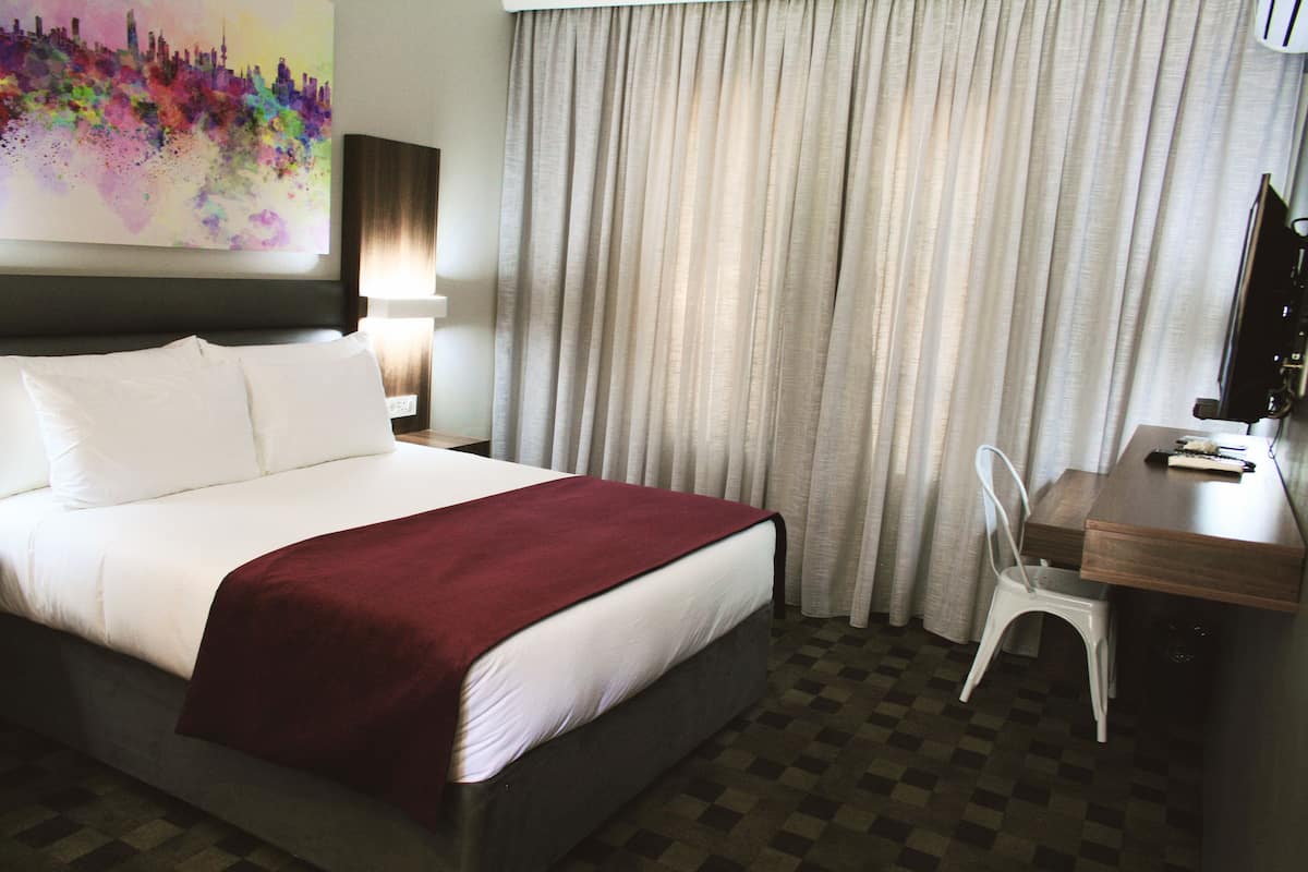 Quarto de hotel com cama, escrivaninha, cadeira, luminária e um quadro na parede.