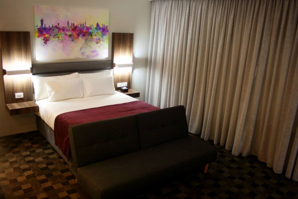 Quarto de hotel com cama, duas mesas de cabeceira, um criado-mudo, sofá, janela grande com cortinas e um quadro abstrato colorido na parede.