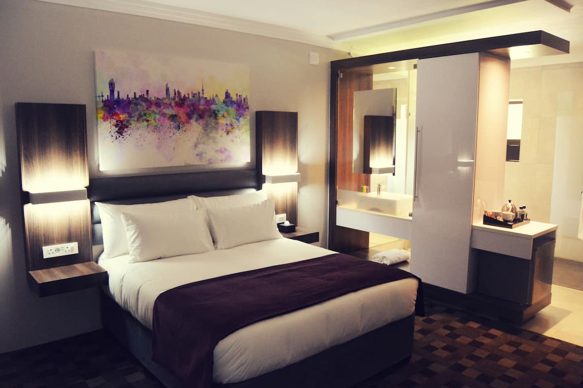 Quarto de hotel com uma cama grande, duas mesas de cabeceira, um criado-mudo, cômoda, espelho e uma obra de arte na parede.