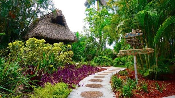 A garden pathway with a thatched-roof structure and a wooden signpost. - Riverside by the Beach Apartments (Pompano Beach)