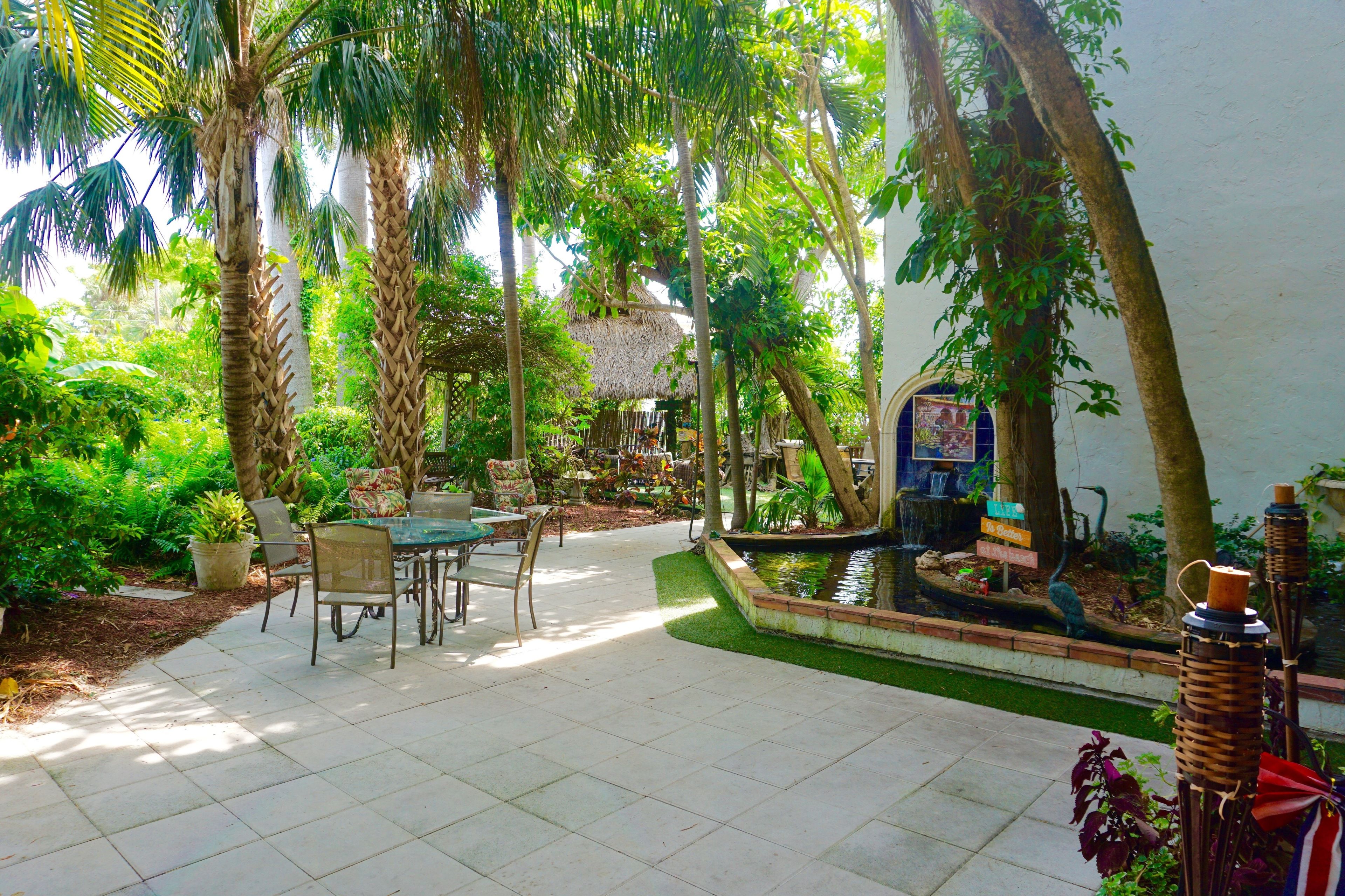 Un patio exterior con una mesa y sillas para comer, rodeado de abundante follaje y una pequeña fuente de agua.
