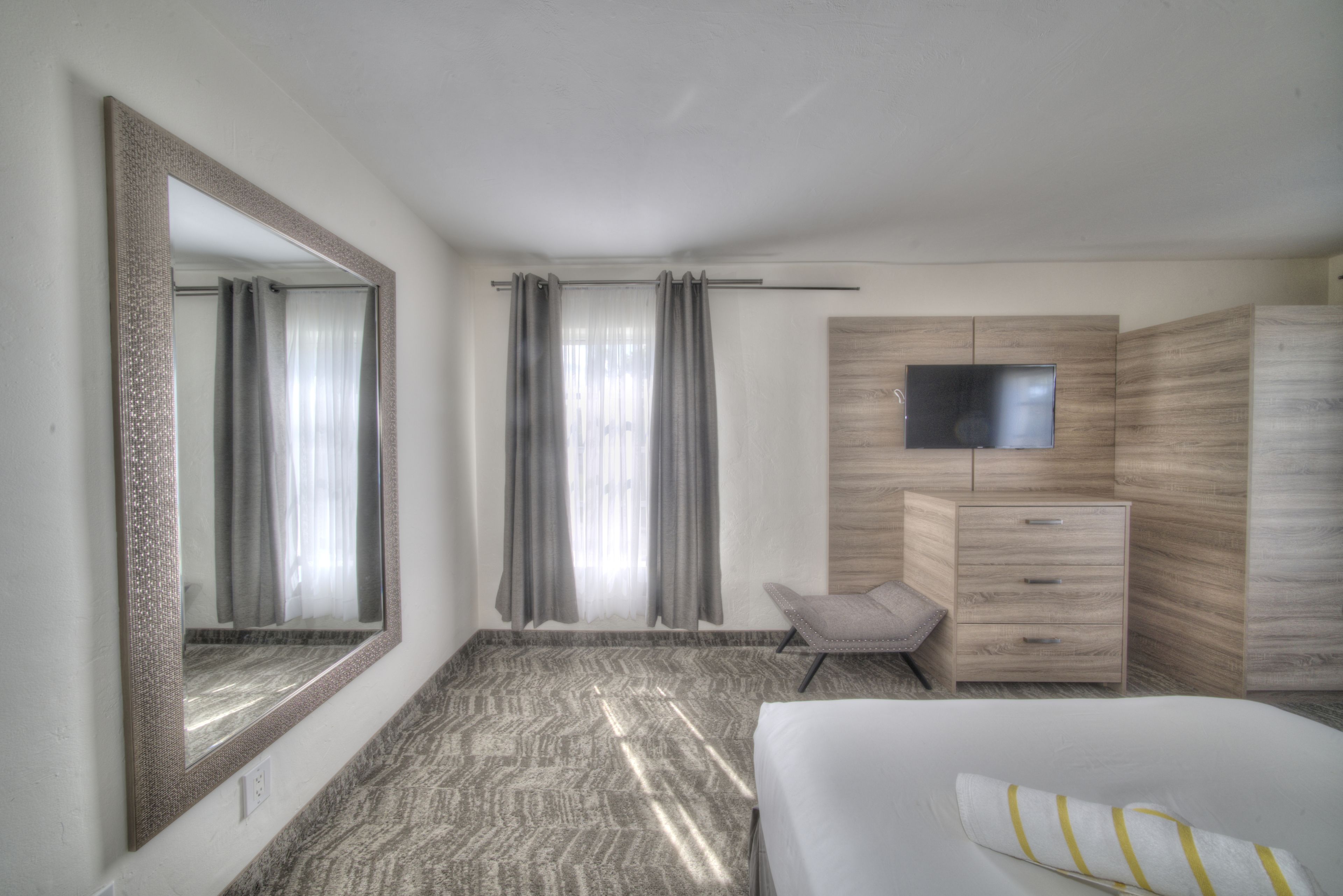A hotel room with a large mirror, a bed, a TV mounted on the wall, a wooden wardrobe, and a chair.