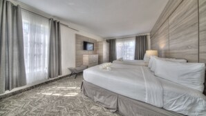A hotel room with a large bed, a flat-screen TV mounted on the wall, a desk with a chair, and a window with curtains. - Riverside by the Beach Apartments (Pompano Beach)