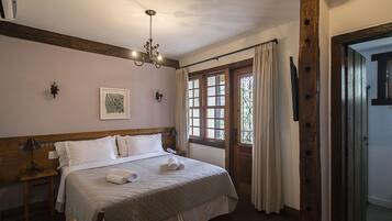 Ein Schlafzimmer mit hölzernem Kopfteil, einem Bett mit weißer Bettwäsche, einer Chandelüster, einem Fenster mit Vorhängen und einer Tür mit Holzrahmen.
