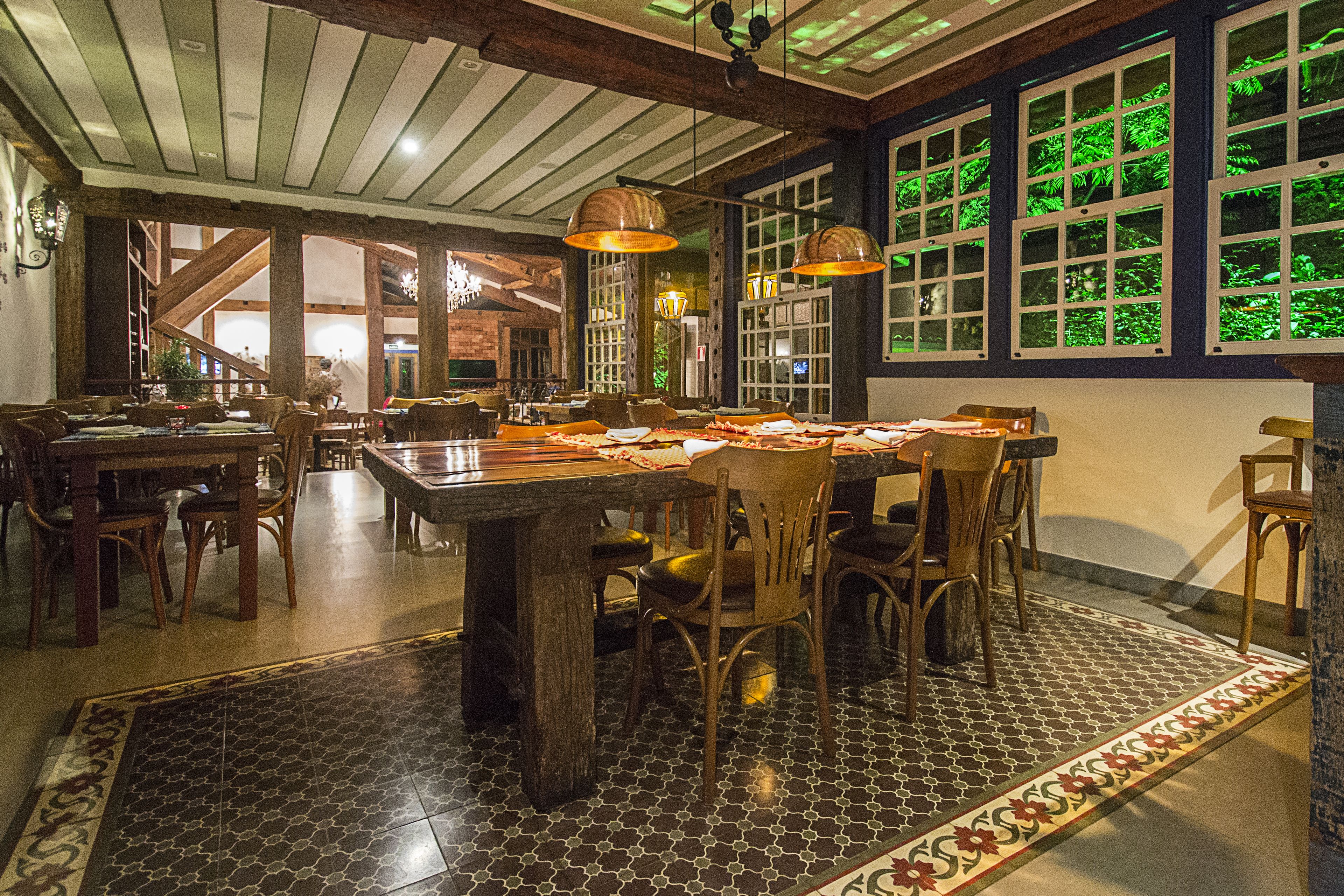 Un comedor con una gran mesa de madera rodeada de sillas, un suelo estampado y ventanas amplias que dan a follaje verde en el exterior.