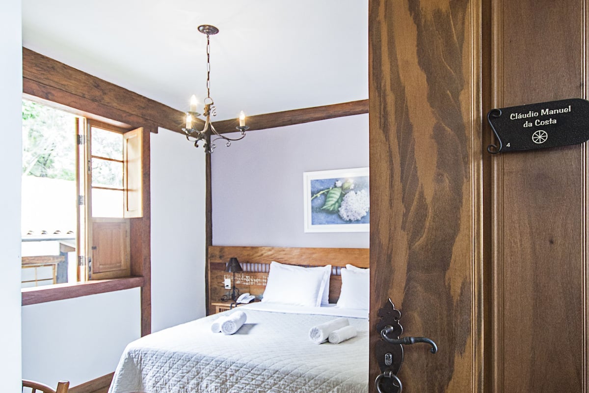 Une chambre à coucher avec une tête de lit en bois, un lit recouvert de draps blancs, un lustre et une porte en bois avec une plaque nominative.