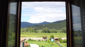 Vista desde una ventana sobre un paisaje verde con colinas, cielo despejado y un pequeño edificio con techo rojo.