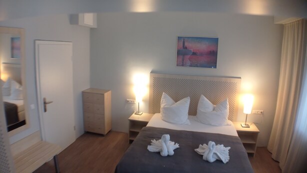 Une chambre d’hôtel avec un lit, des lampes de chevet, une table de chevet, une commode, un banc, un tableau au mur et une porte.