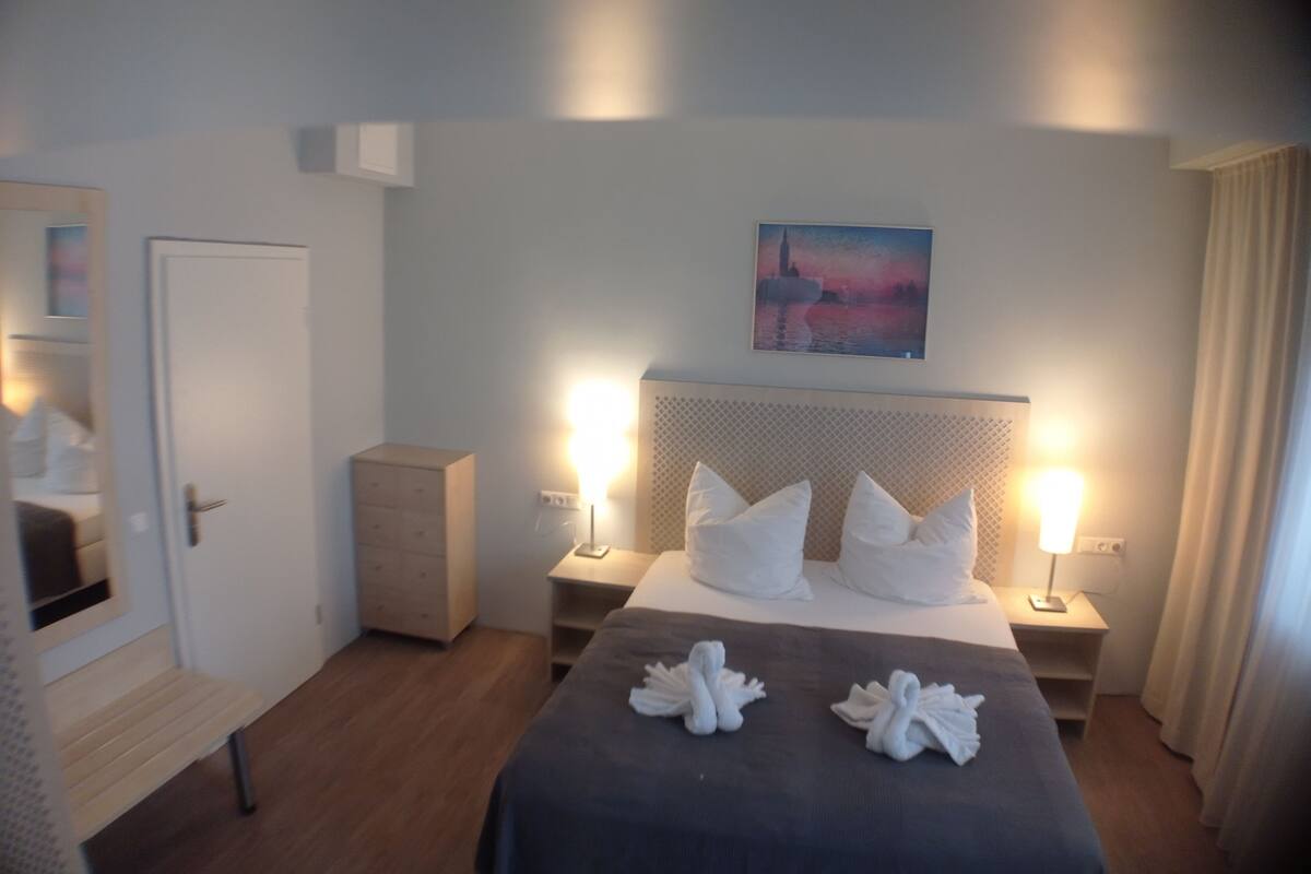 Una camera d'albergo con un letto, abat-jour, comodino, una cassettiera, una panca, un quadro appeso al muro e una porta.
