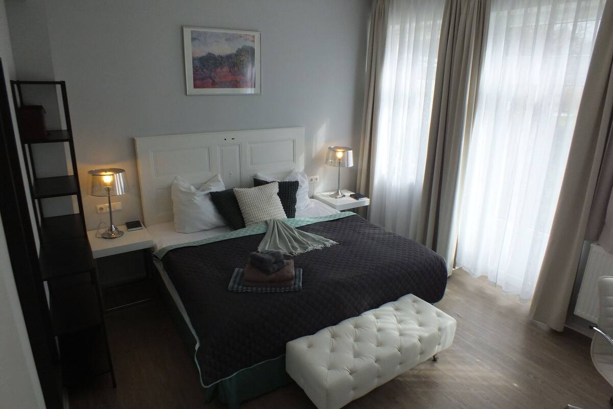 Una camera d'albergo ordinata con un letto, comodini, una lampada, una sedia e un quadro appeso alla parete.