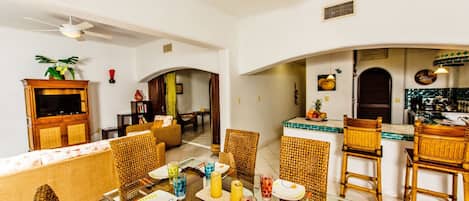 A spacious living area with a dining table set for four, surrounded by wicker chairs. A kitchen area with a bar counter and stools is visible in the background.