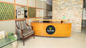 A reception desk with a wooden finish and the logo "Ayenda Hotels". There is a striped armchair to the left of the desk. - Hotel  Oasis (Rionegro)