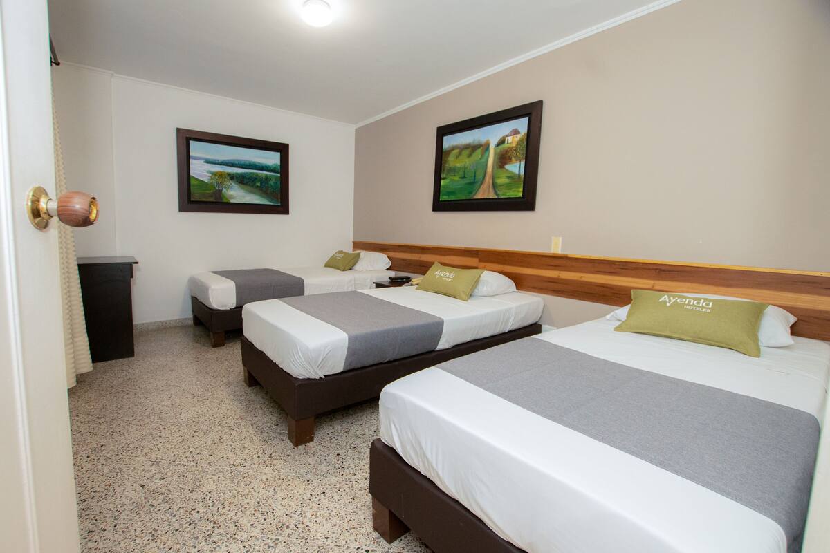 Hotelkamer met drie bedden, elk voorzien van een houten hoofdbord, wit beddengoed en een groen kussen met de naam van het hotel. Twee ingelijste foto’s hangen aan de muur, en de kamer heeft een vloerbedekking.