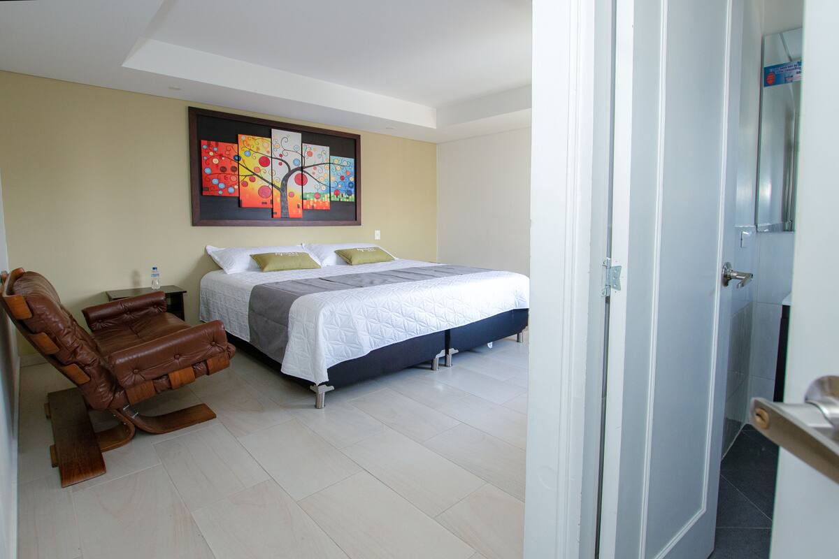 Een slaapkamer met een bed, een fauteuil, een schilderij aan de muur en een deur naar een andere kamer.
