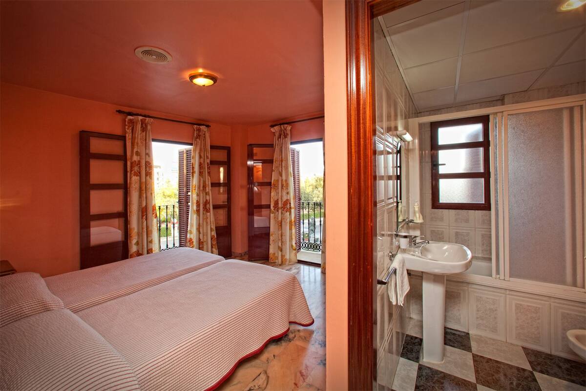 A room with a bed, a bathroom with a sink and mirror, and a view through a window with curtains.