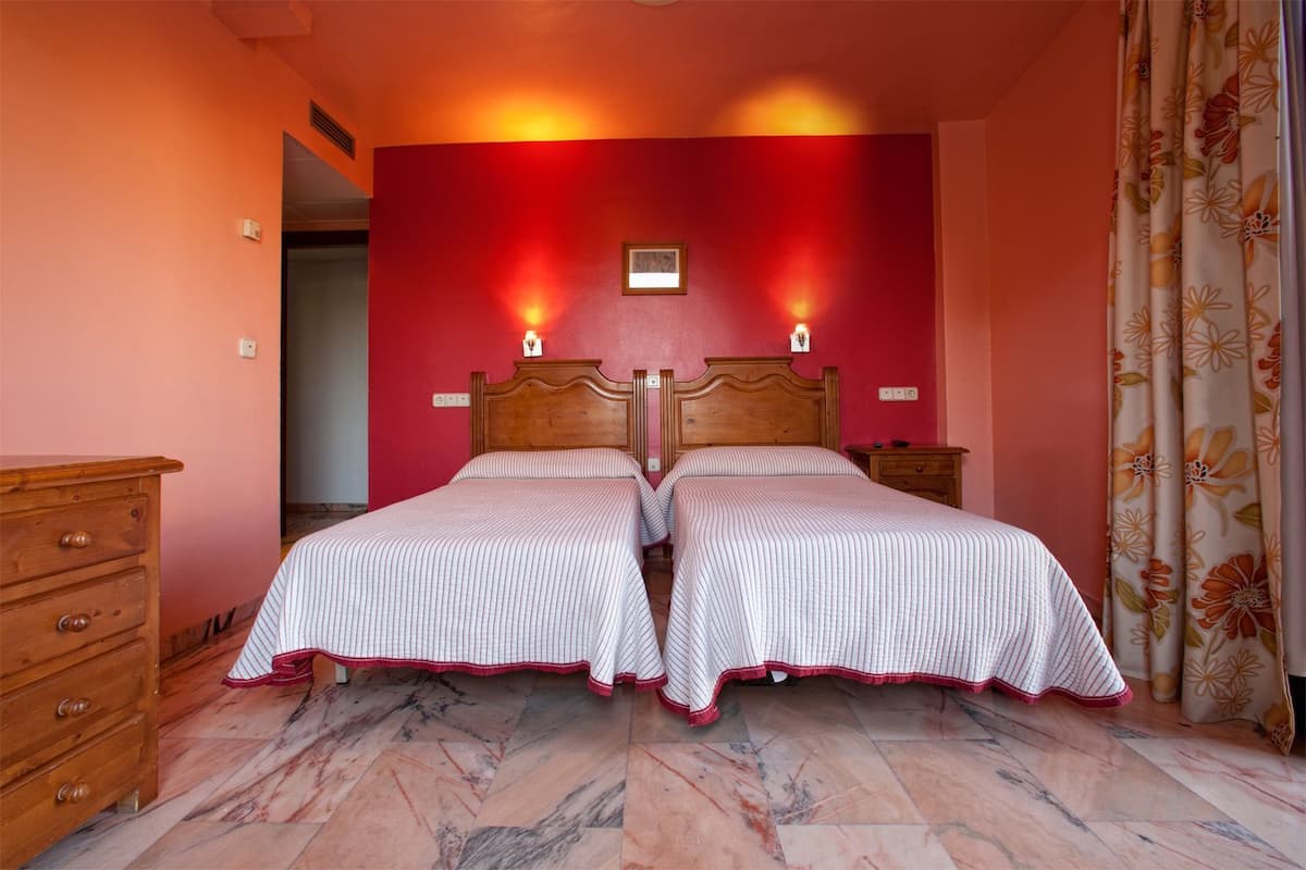 A double bed with a wooden headboard, two bedside tables, a wooden dresser, and a red accent wall.