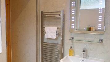 A bathroom with a glass shower enclosure, a towel rack with a white towel, a mirror with lights, and a sink with toiletries.