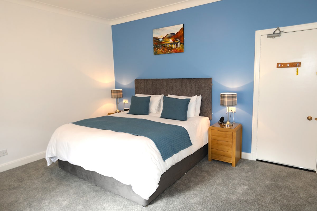 A bedroom with a bed, bedside tables, a nightstand, a door, and a wall-mounted painting.