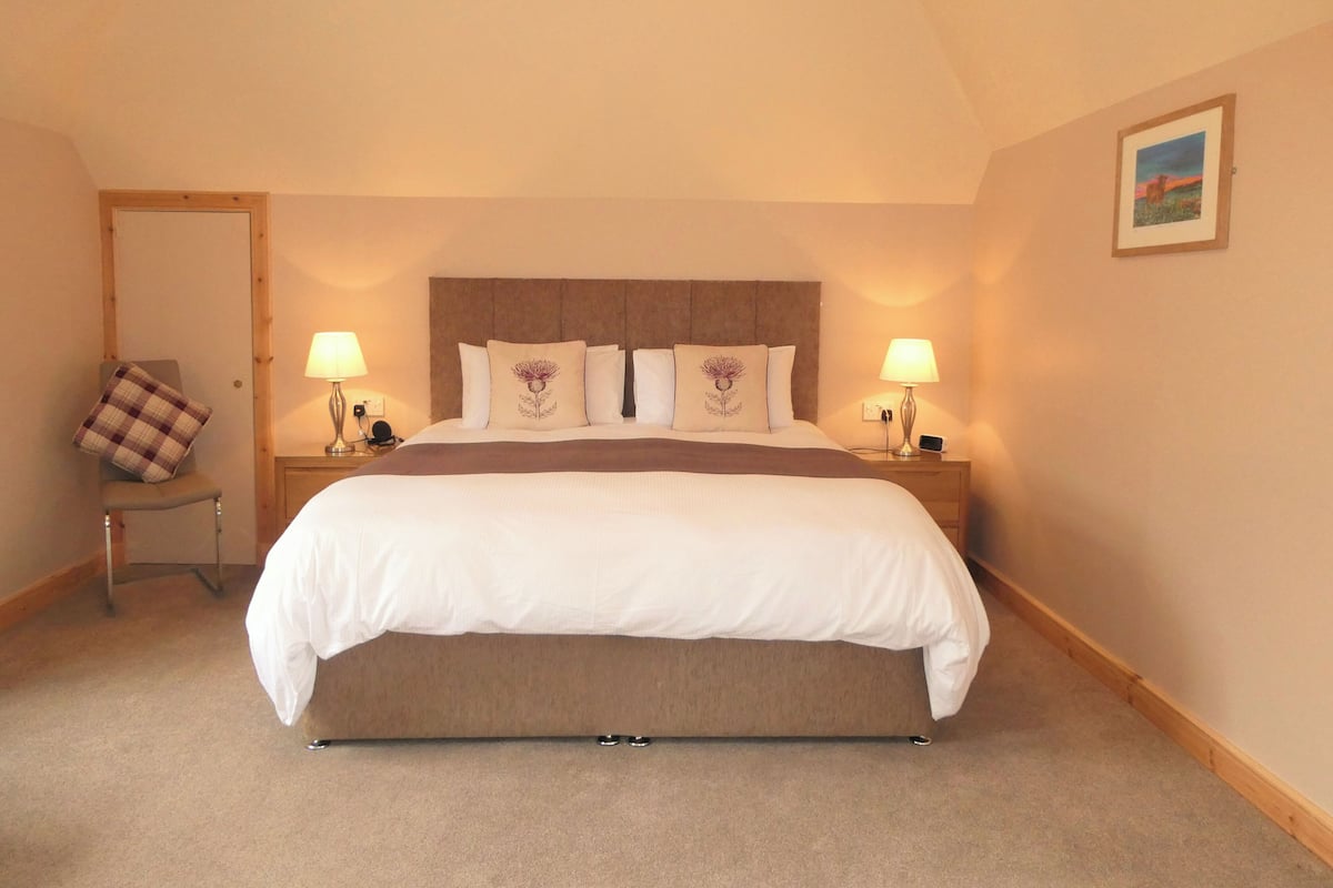 A neatly made bed with a brown headboard, two bedside lamps, and a chair with a plaid cushion.