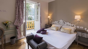 Un dormitorio con cama, lámparas de noche, una silla, una mesita de vidrio, una ventana con cortinas y vistas a palmeras.