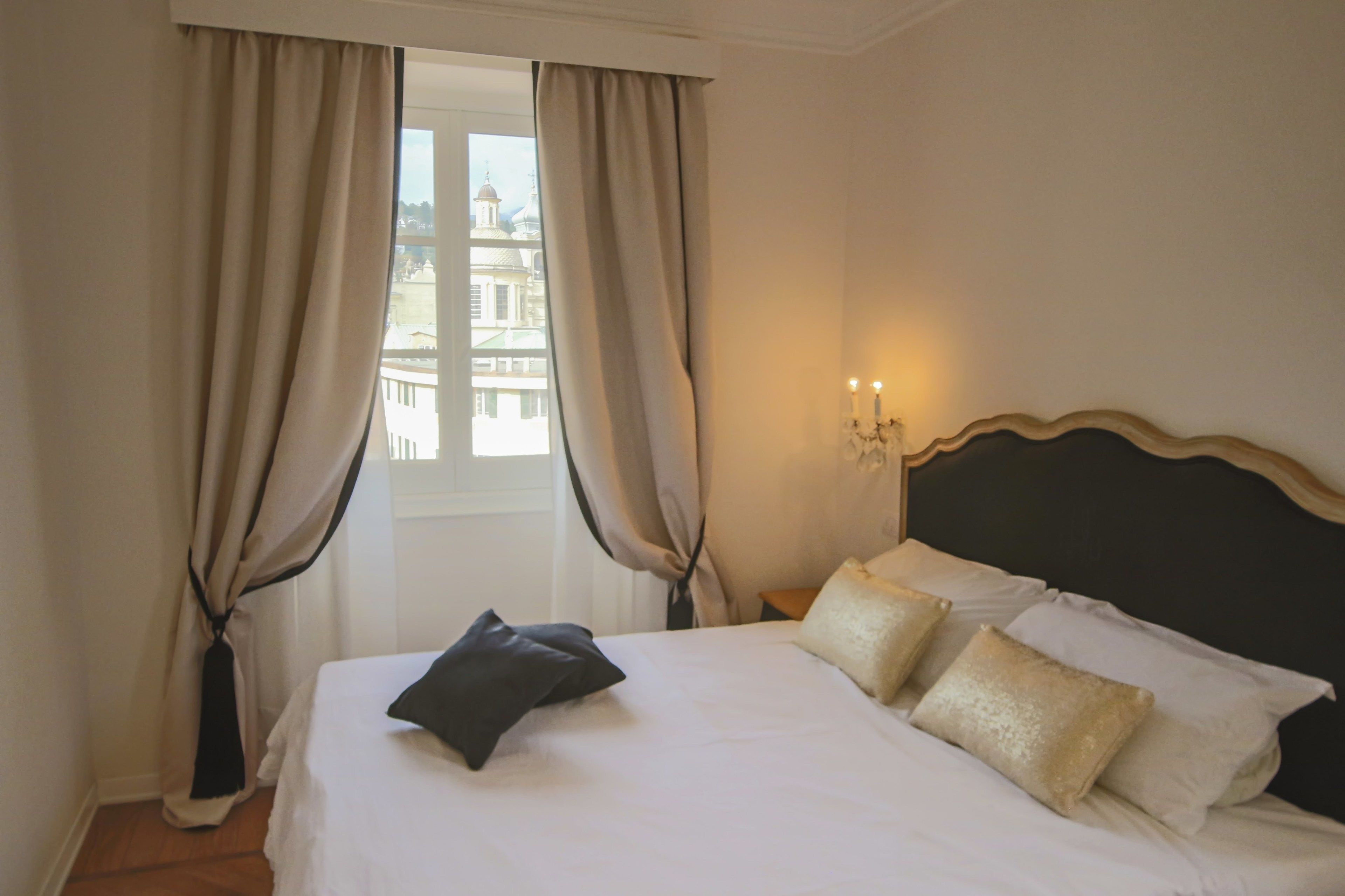 A hotel room with a bed, beige curtains, a view of a building, and a wall-mounted light fixture.