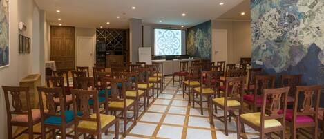 A conference room with wooden chairs arranged in rows, a projector screen, and a large abstract painting on the wall.