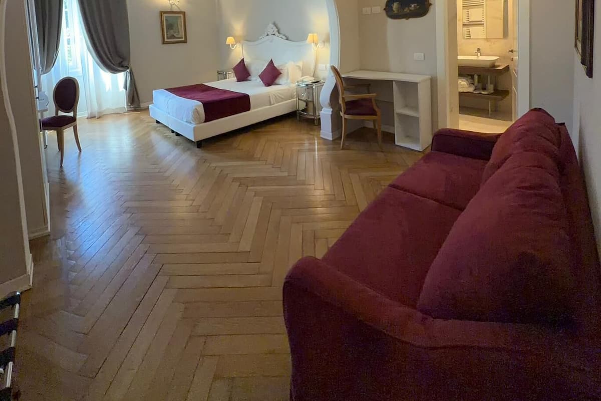 A hotel room with a bed, a red sofa, a desk, and a bathroom visible through an archway.