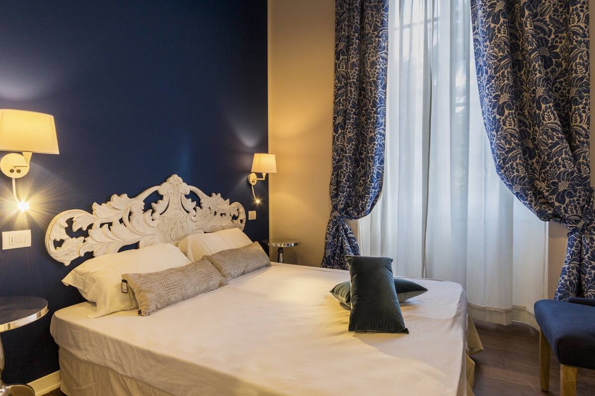 A hotel room with a bed, two bedside lamps, a patterned curtain, and a chair.