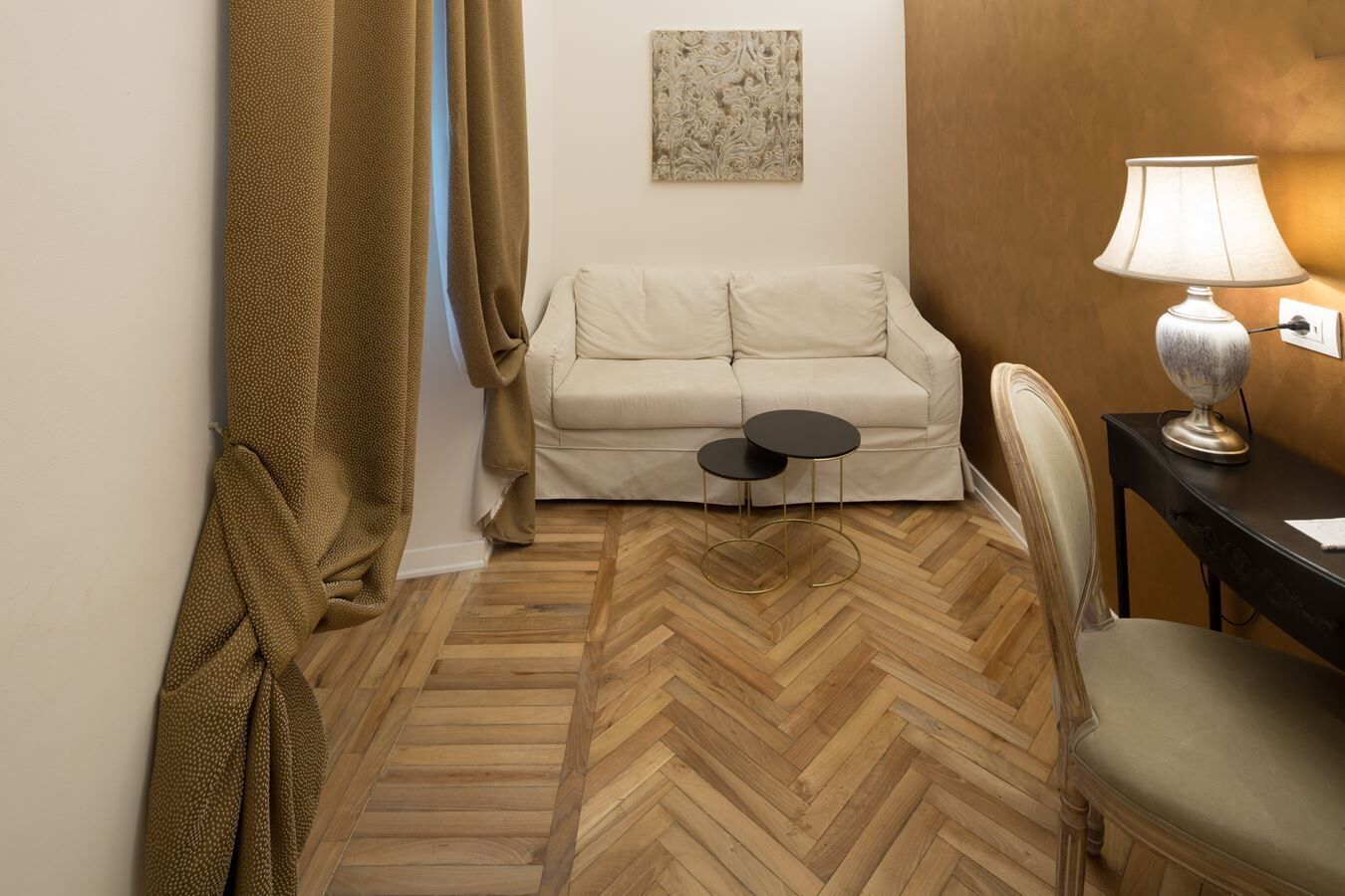 Une chambre d’hôtel avec un parquet en bois à motifs chevrons, un canapé beige, une petite table ronde, une lampe et une chaise.